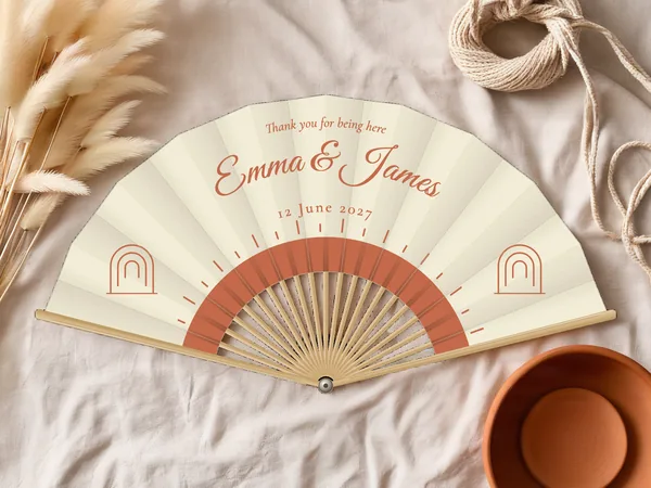 Personalised wedding fan "Boho Sun" with the names Emma & James, a radiant half sun below and small arches on the sides, ivory fabric with terracotta print, on a wedding table