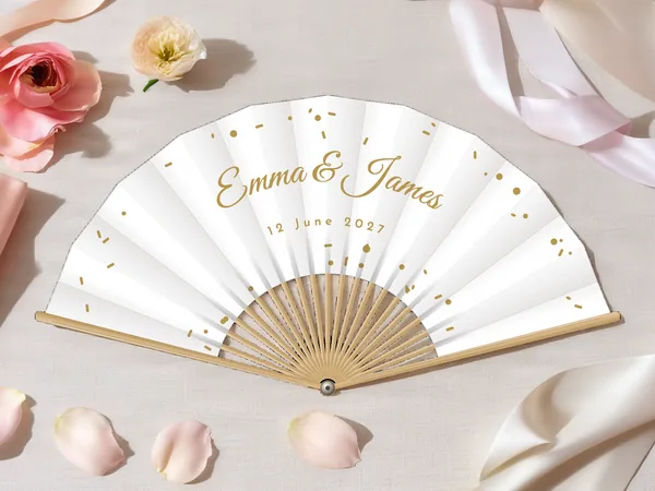 Personalised wedding fan "Confetti" with the names Emma & James, tiny confetti scattered all around, white fabric with gold print, on a wedding table