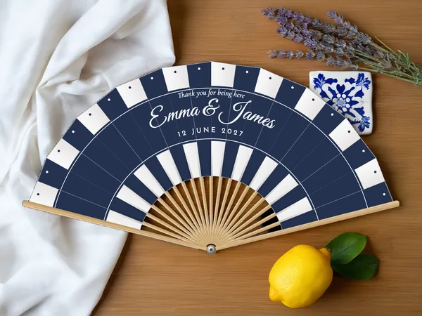Personalised wedding fan "Deckchair Stripes" with the names Emma & James, parasol stripes in bands along the edge and the bottom, white fabric with navy blue print, on a wedding table