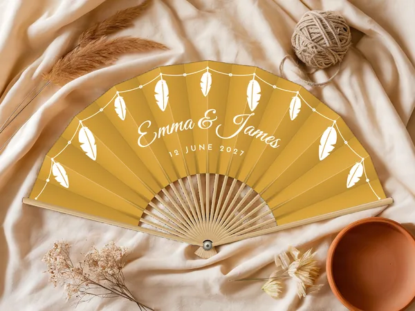 Personalised wedding fan "Feathers" with the names Emma & James, feathers hanging from a beaded garland along the edge, mustard fabric with white print, on a wedding table