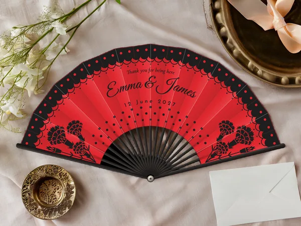 Personalised wedding fan "Flamenco" with the names Emma & James, a polka-dot lace ruffle along the edge and carnations in the corners, red fabric with black print, on a wedding table
