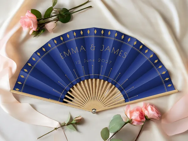 Personalised wedding fan "Gatsby" with the names Emma & James, diamonds along the edge and Art Deco rays below, navy blue fabric with gold print, on a wedding table