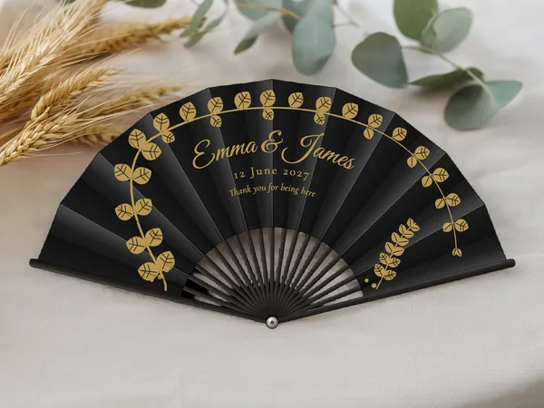 Personalised wedding fan "Gilded Eucalyptus" with the names Emma & James, arching eucalyptus branches framing them, black fabric with gold print, on a wedding table