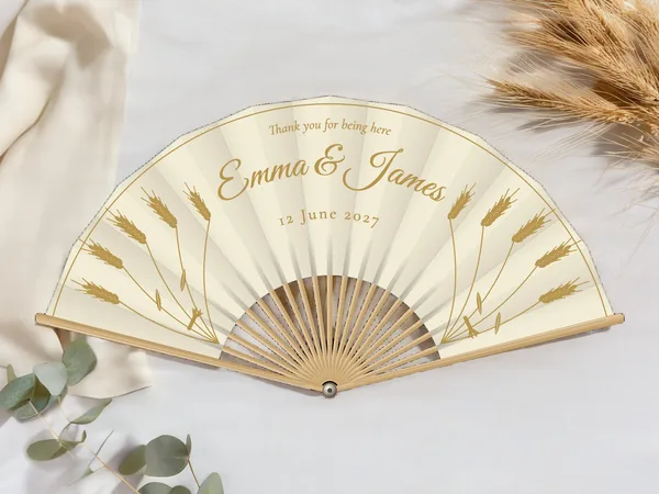 Personalised wedding fan "Golden Wheat" with the names Emma & James, upright wheat stalks on each side and a fine border line, ivory fabric with gold print, on a wedding table