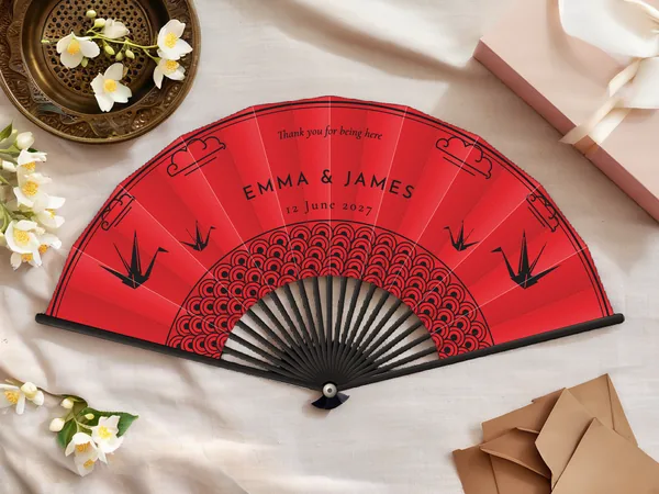 Personalised wedding fan "Kimono" with the names Emma & James, origami cranes, clouds and a wave pattern around the centre, red fabric with black print, on a wedding table