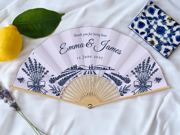 Personalised wedding fan "Lavender Fields" with the names Emma & James, lavender bunches, bees and a lavender field beneath, lavender fabric with navy blue print, on a wedding table