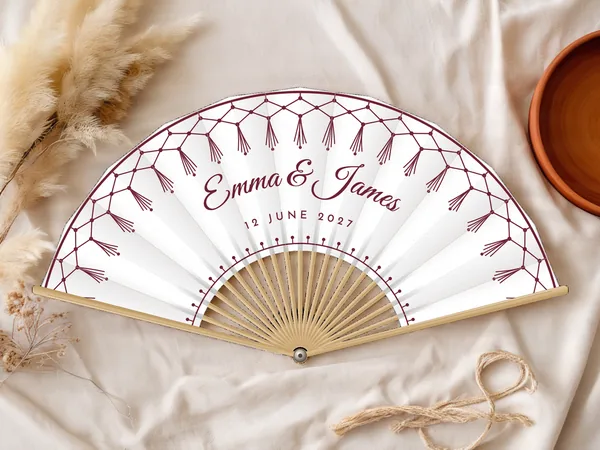 Personalised wedding fan "Macramé" with the names Emma & James, a tasselled macramé fringe along the edge, white fabric with burgundy print, on a wedding table