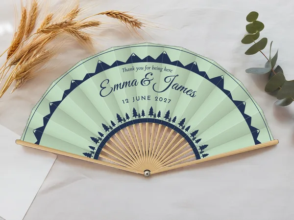 Personalised wedding fan "Pines and Peaks" with the names Emma & James, mountain ridges along the edge and a row of pine trees at the bottom, seafoam fabric with navy blue print, on a wedding table