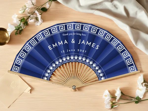 Personalised wedding fan "Santorini" with the names Emma & James, a Greek key border and a wreath of flowers around the centre, navy blue fabric with white print, on a wedding table