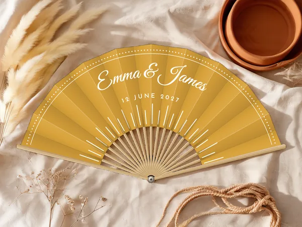Personalised wedding fan "Sunbeams" with the names Emma & James, fine rays rising from the bottom and a dotted border, mustard fabric with white print, on a wedding table