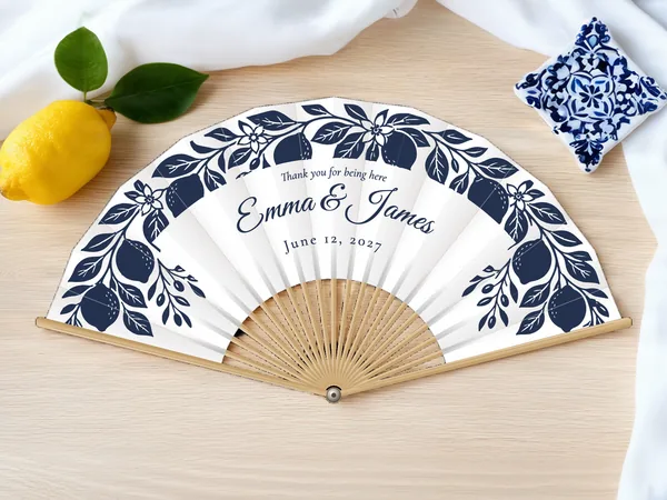 Personalized wedding fan "Amalfi Lemons" with the names Emma & James, lemon branches and blossoms framing them, white fabric with navy blue print, on a wedding table