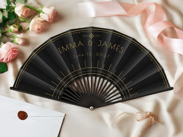 Personalized wedding fan "Art Deco" with the names Emma & James, fine arcs and graphic Art Deco rays around the center, black fabric with gold print, on a wedding table