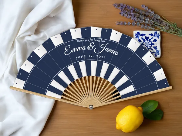 Personalized wedding fan "Cabana Stripes" with the names Emma & James, beach umbrella stripes in bands along the edge and the bottom, white fabric with navy blue print, on a wedding table