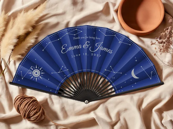 Personalized wedding fan "Celestial" with the names Emma & James, constellations, a sun, and a crescent moon scattered all around, navy blue fabric with silver print, on a wedding table