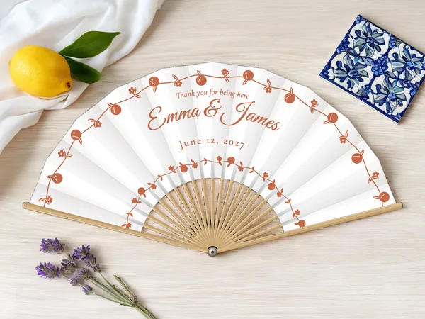 Personalized wedding fan "Dolce Vita" with the names Emma & James, arching garlands of little citrus fruits and flowers all around, white fabric with terracotta print, on a wedding table