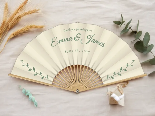 Personalized wedding fan "Eucalyptus" with the names Emma & James, slender leafy branches on either side below, ivory fabric with sage green print, on a wedding table