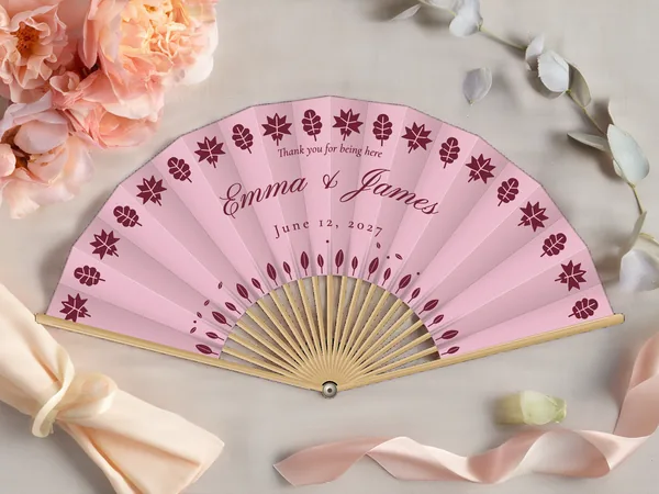Personalized wedding fan "Fall Leaves" with the names Emma & James, a frieze of stylized leaves all the way around, blush pink fabric with burgundy print, on a wedding table