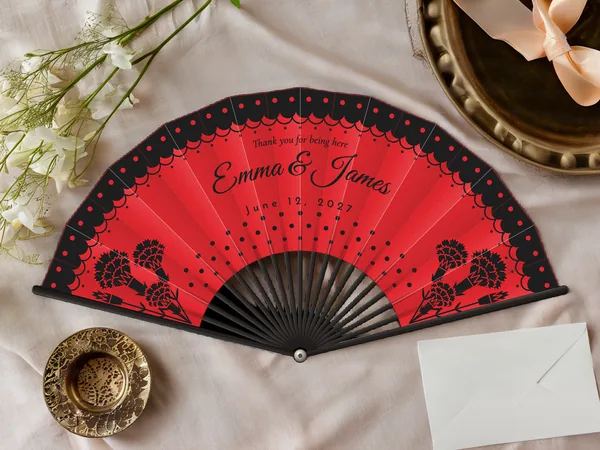 Personalized wedding fan "Flamenco" with the names Emma & James, a polka-dot lace ruffle along the edge and carnations in the corners, red fabric with black print, on a wedding table