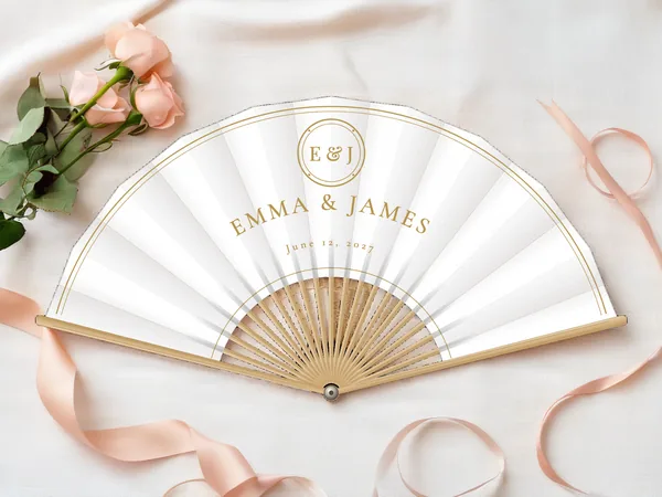Personalized wedding fan "Gold Monogram" with the names Emma & James, an initial medallion above and a double line along the edge, white fabric with gold print, on a wedding table