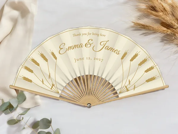 Personalized wedding fan "Golden Wheat" with the names Emma & James, upright wheat stalks on each side and a fine border line, ivory fabric with gold print, on a wedding table
