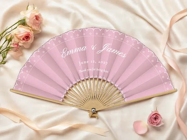 Personalized wedding fan "Lace" with the names Emma & James, a scalloped lace-style border and dotted lines along the edge, blush pink fabric with white print, on a wedding table