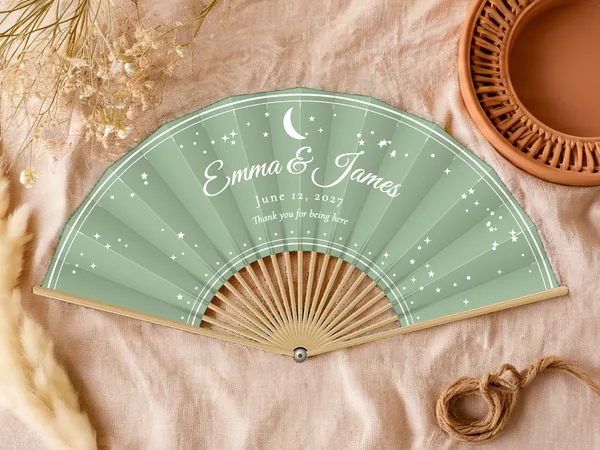 Personalized wedding fan "Moon and Stars" with the names Emma & James, a crescent moon and scattered stars all around, sage green fabric with white print, on a wedding table