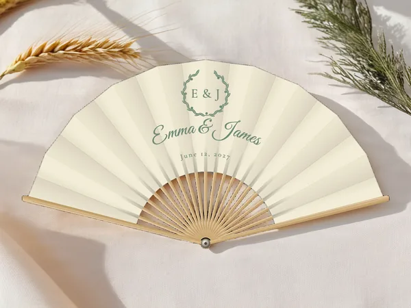 Personalized wedding fan "Olive Wreath" with the names Emma & James, a leafy wreath circling the initials above, ivory fabric with sage green print, on a wedding table