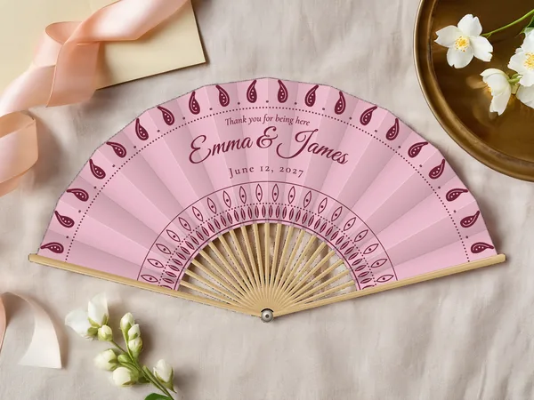 Personalized wedding fan "Paisley" with the names Emma & James, paisley drops along the edge and a rosette around the center, blush pink fabric with burgundy print, on a wedding table
