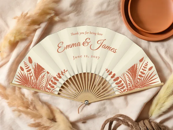 Personalized wedding fan "Pampas Grass" with the names Emma & James, pampas grass, palm fronds, and dried foliage in the lower corners, ivory fabric with terracotta print, on a wedding table