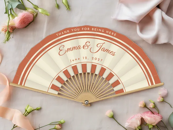 Personalized wedding fan "Rainbow" with the names Emma & James, wide arched bands at the edge and a crenellated ring around the center, ivory fabric with terracotta print, on a wedding table