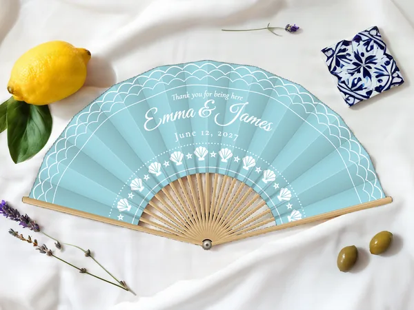 Personalized wedding fan "Seaside" with the names Emma & James, waves along the edge and an arc of seashells below, sky blue fabric with white print, on a wedding table