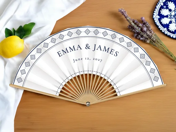 Personalized wedding fan "Spanish Tile" with the names Emma & James, a border of diamond-shaped tiles along the edge, white fabric with navy blue print, on a wedding table
