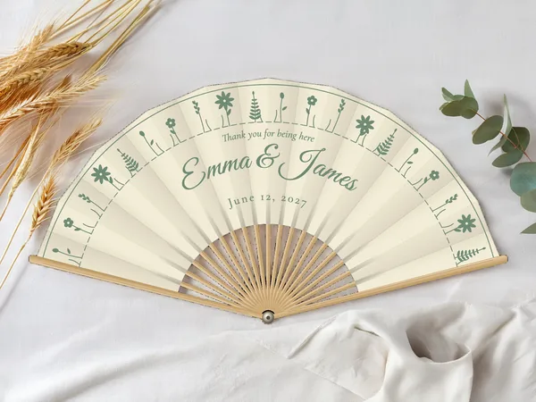 Personalized wedding fan "Wildflowers" with the names Emma & James, a ring of tiny wildflowers all around, ivory fabric with sage green print, on a wedding table